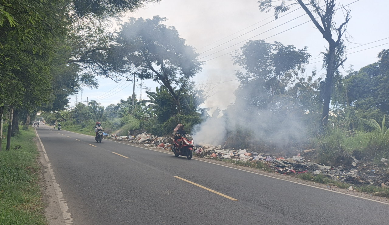 Tumpukan sampah terlihat dibakar di pinggir jalan raya, menimbulkan asap tebal yang mengganggu pengendara dan mencemari lingkungan sekitar)Bn.